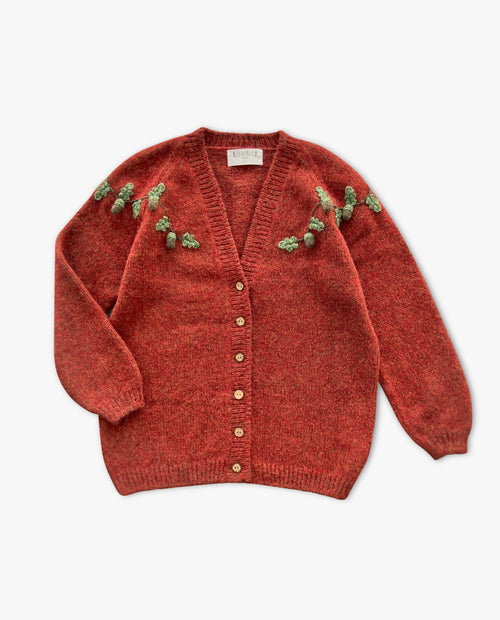 Red cardigan with button details and green floral embellishments on a white background